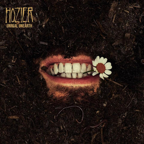 a square of dirt/mulch. in the center, a mouth is smiling and biting a white-petaled flower's stem. in the top left corner, beige-yellow font reads "HOZIER" in all-caps. below it, in smaller text: "UNREAL UNEARTH" also in all-caps.
